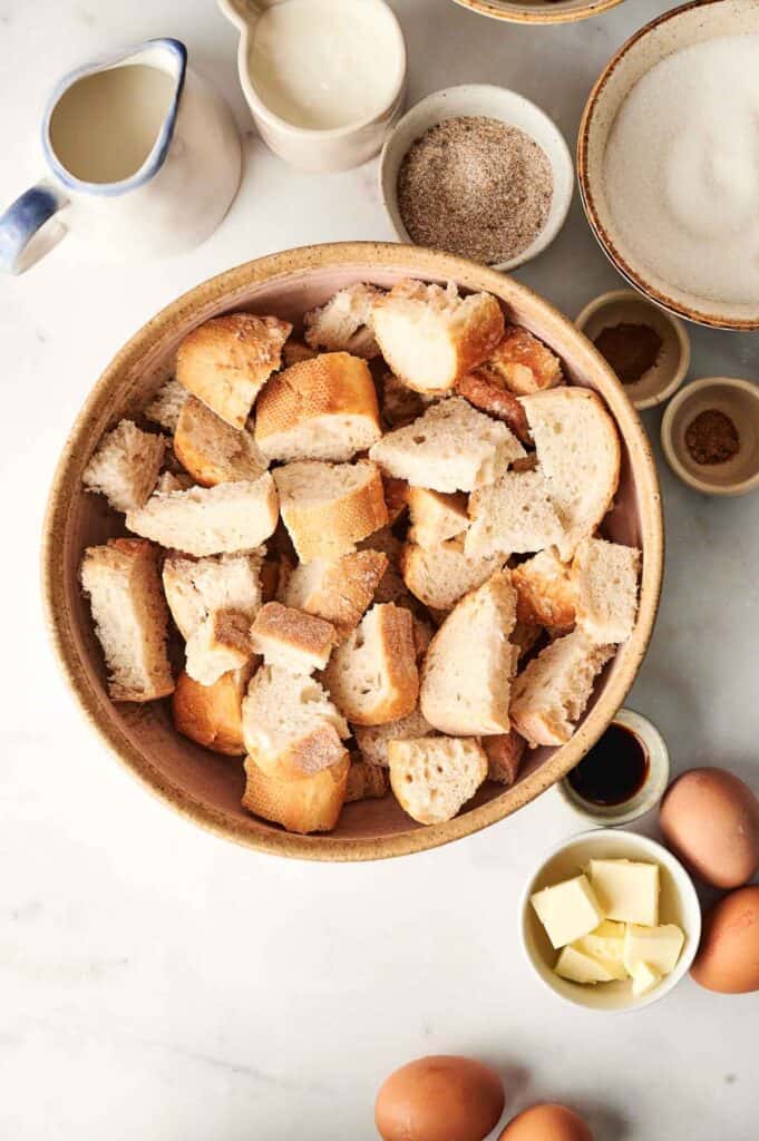 A bowl of bread cubes is surrounded by a jug of milk, cream, sugar, spices, butter, whole eggs, and vanilla on a marble surface&mdash;perfect for crafting an air fryer bread pudding.
