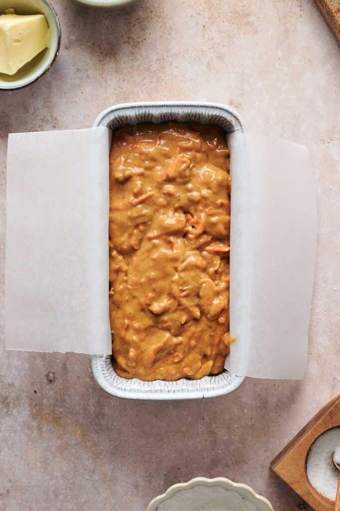 Batter with visible ingredients, reminiscent of a rich carrot cake, spread in a loaf pan lined with parchment paper on a textured surface. Nearby, bowls with butter and spices add to the enticing scene.