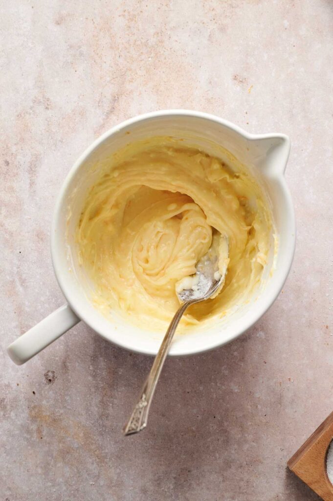 A mixing bowl with creamy yellow carrot cake batter and a metal spoon resting inside on a light textured surface.