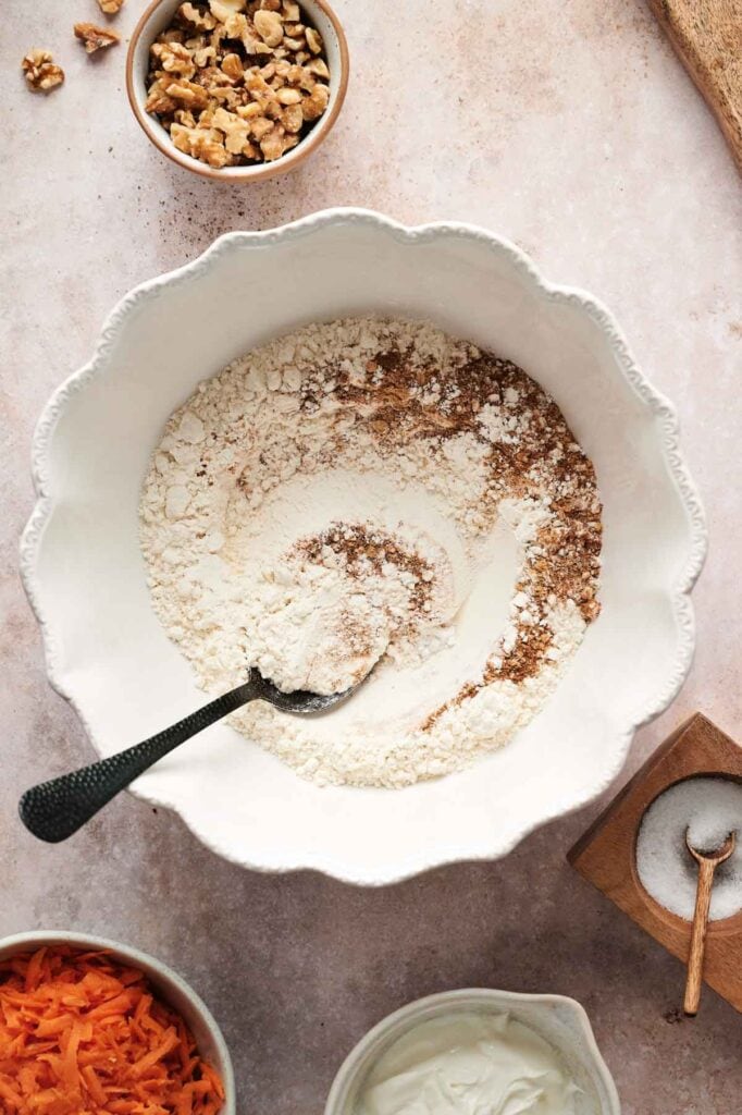 A mixing bowl with flour, spices, and a spoon awaits the magic touch of creating carrot cake. Surrounding it are bowls of walnuts, shredded carrots, yogurt, and a wooden spoon with salt.