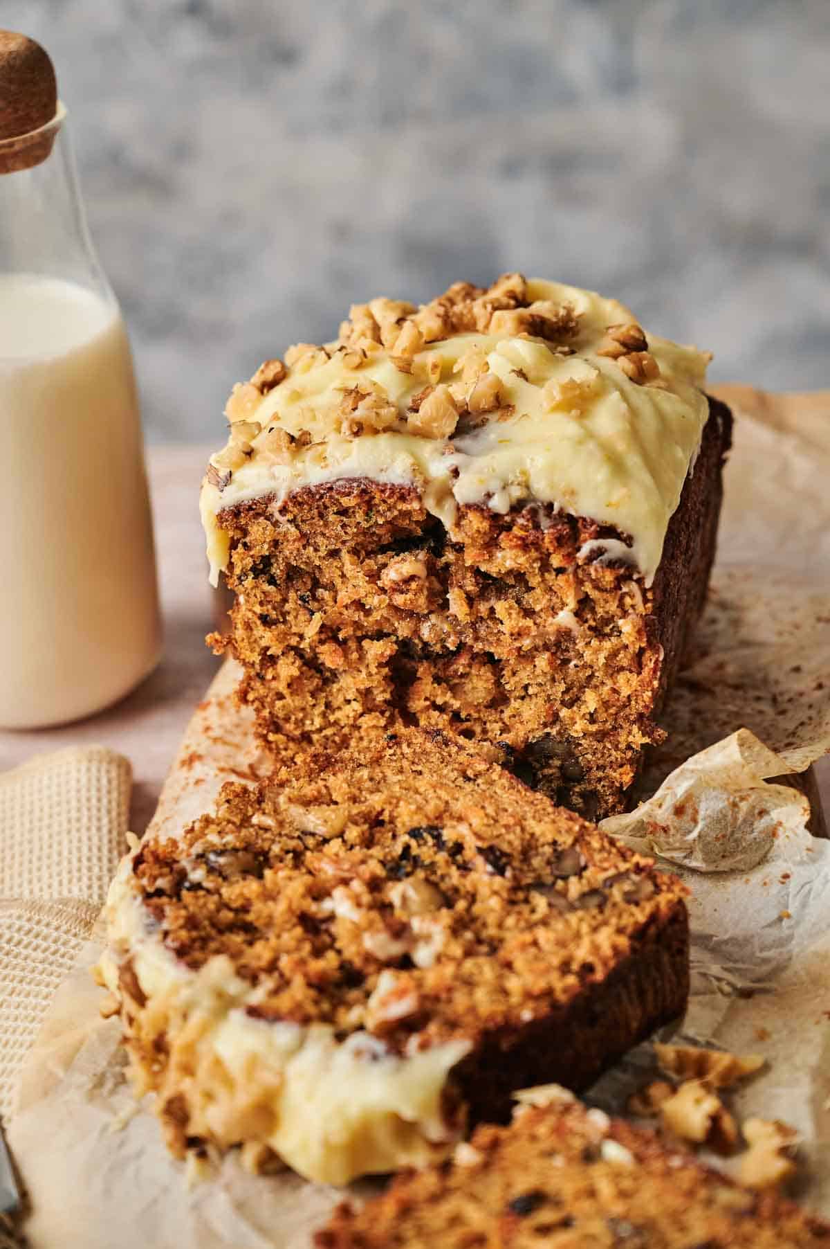 A scrumptious loaf of carrot cake, adorned with cream cheese frosting and sprinkled with nuts, rests on parchment paper, partially sliced. A bottle of milk stands invitingly in the background.