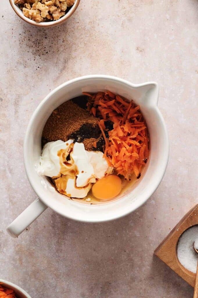 A mixing bowl with shredded carrots, egg, yogurt, brown sugar, vanilla, and cinnamon on a marble surface is the perfect start of a carrot cake. Nearby are small bowls filled with walnuts and spices.