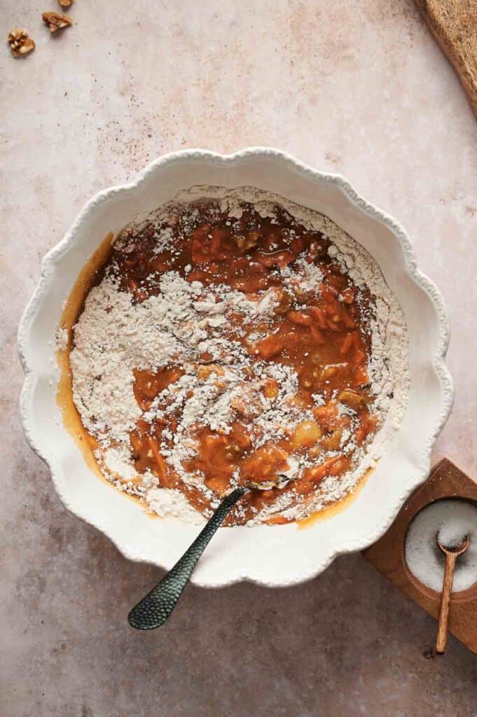 A bowl filled with mixed ingredients, hinting at carrot cake prep, includes flour and a brown liquid, with a spoon resting inside. Nearby sits a small wooden container holding salt, complete with its own spoon.