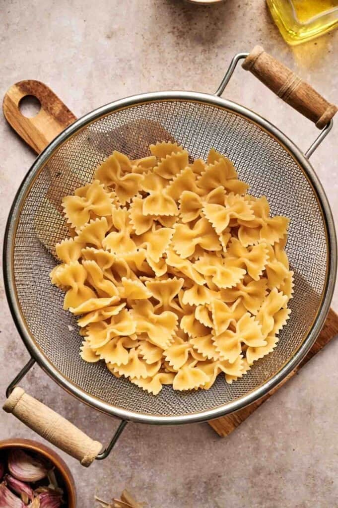 Cooked farfalle pasta, perfect for chicken Alfredo, rests in a metal colander on a wooden cutting board.