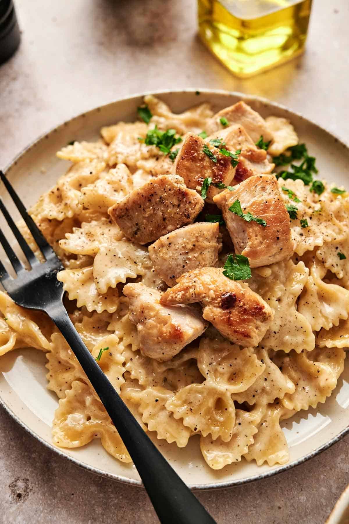 Plate of bowtie pasta with creamy Alfredo sauce, topped with grilled chicken pieces and garnished with parsley; a black fork rests on the plate.