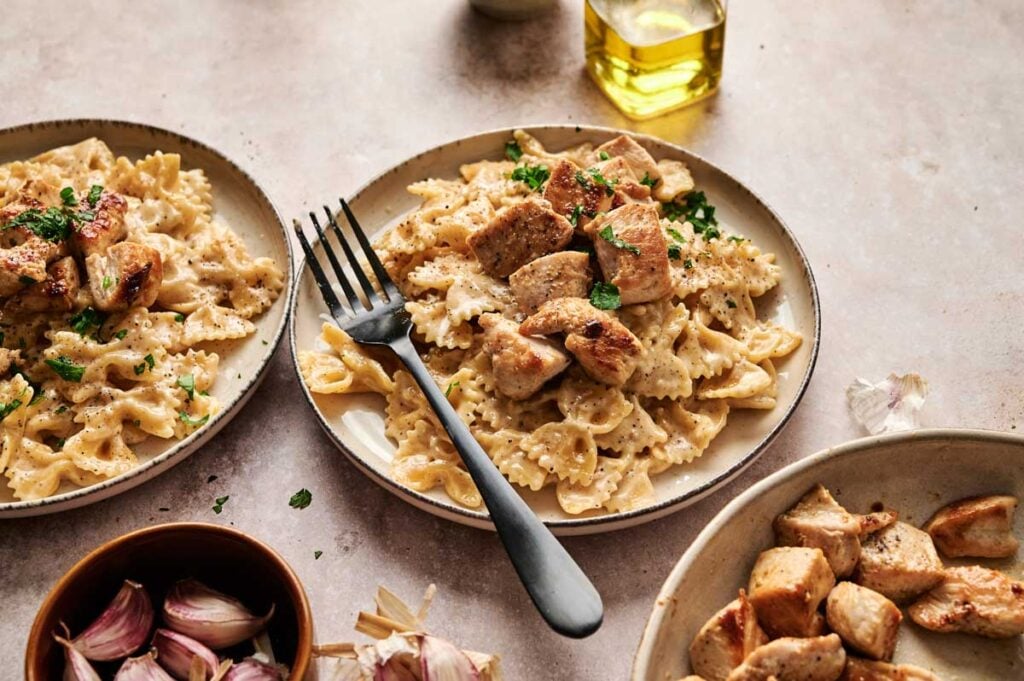 Plate of bowtie pasta with creamy chicken alfredo sauce and grilled chicken pieces, garnished with herbs. A fork rests on the plate; garlic, chicken, and olive oil are nearby.