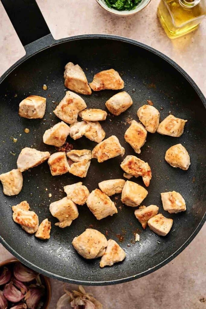 Chunks of browned chicken sizzle in a black frying pan, preparing the perfect base for a delectable chicken Alfredo, with garlic and other fresh ingredients scattered around.