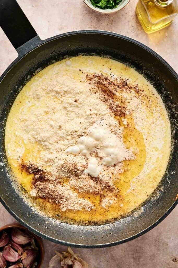 A skillet filled with creamy chicken Alfredo sauce is being mixed with grated cheese and spices. Garlic, parsley, and olive oil are visible nearby.