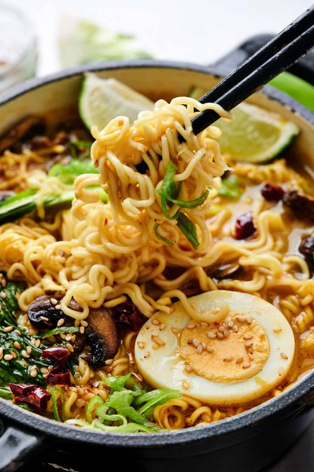 A bowl of ramen with noodles, sliced egg, mushrooms, greens, and sesame seeds, held with chopsticks. Lime wedges are in the background.