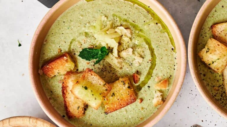 A bowl of creamy broccoli soup topped with croutons, shredded cheese, a mint leaf, and a drizzle of olive oil.