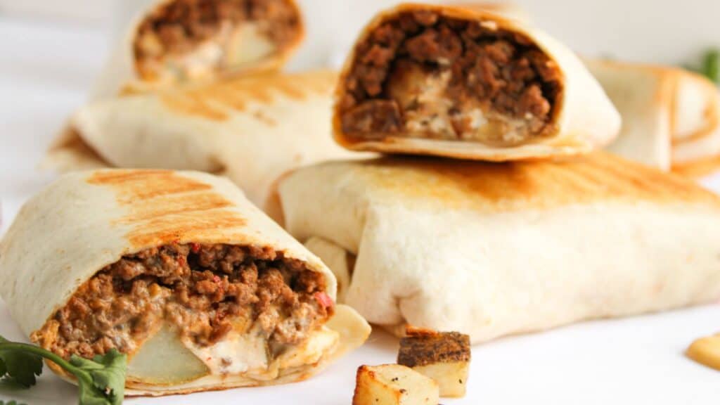 Close-up of several wrapped burritos on a table, with some cut open showing a filling of ground meat and sauce. Pieces of roasted potato and cilantro are scattered around.