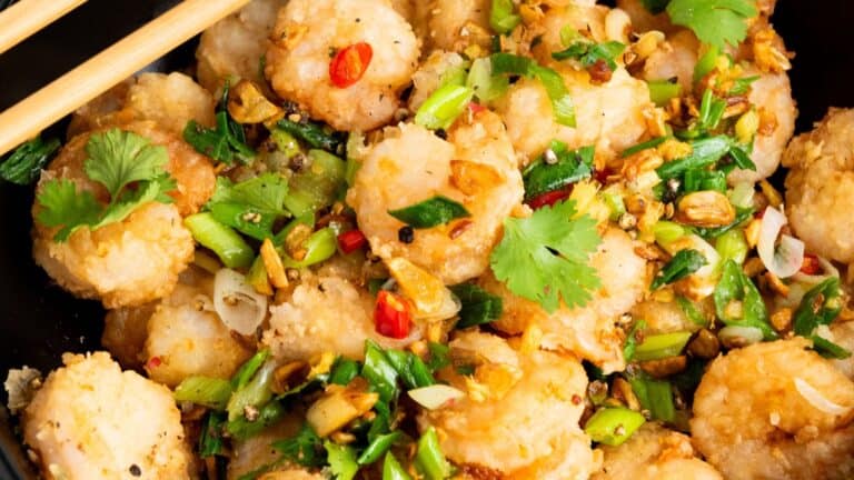 A close-up of a dish featuring fried shrimp garnished with chopped green onions, red chili slices, garlic, and cilantro, with chopsticks resting on the side.