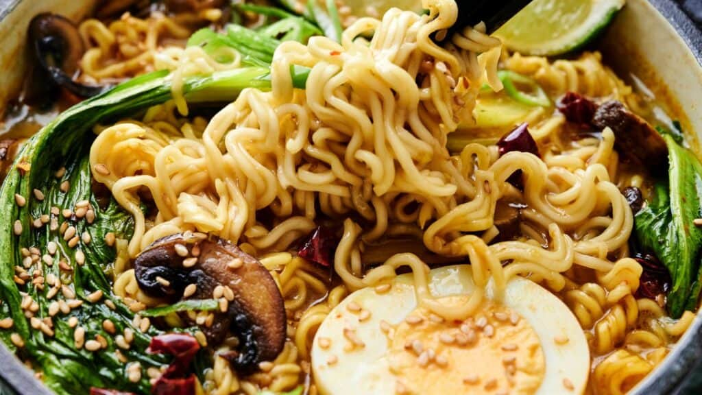 Close-up of a bowl of ramen with noodles, bok choy, mushrooms, half a boiled egg, sesame seeds, and lime, garnished with chili slices. Black chopsticks hold some noodles above the bowl.