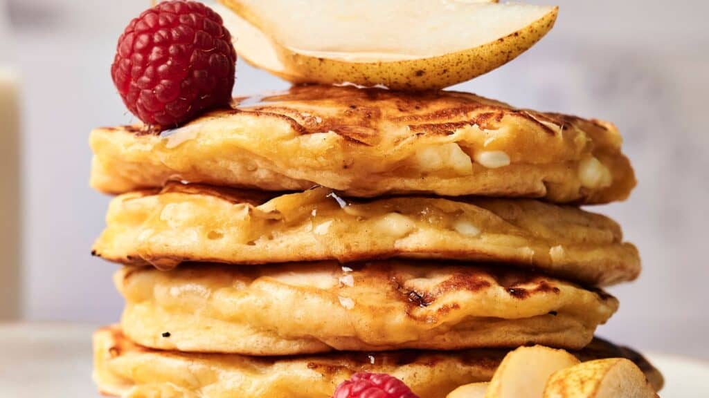 A stack of pancakes topped with a pear slice, raspberry, and syrup drizzle, with almonds and raspberries on the side.