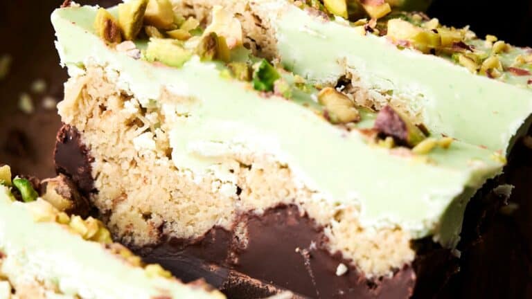 Close-up of sliced pistachio dessert bars with a green topping, chopped pistachios, a crumbly middle layer, and a dark chocolate base.