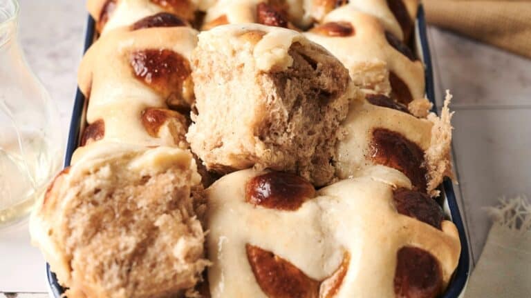 A tray of freshly baked hot cross buns, with shiny tops and cross patterns, one bun partially pulled apart.