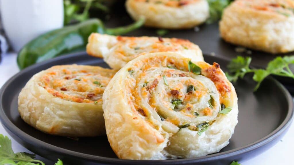 Close-up of puff pastry pinwheels filled with cheese and herbs on a dark plate, with a green chili pepper and cilantro in the background.