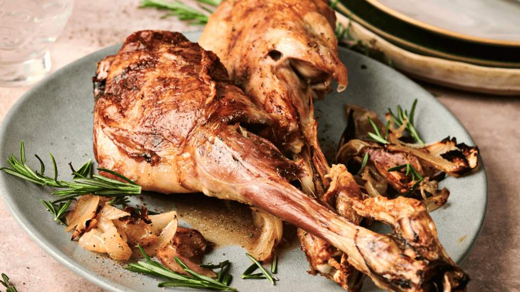 Roasted lamb shanks served on a plate with rosemary and roasted onions, garnished with herbs, accompanied by stacked plates in the background.