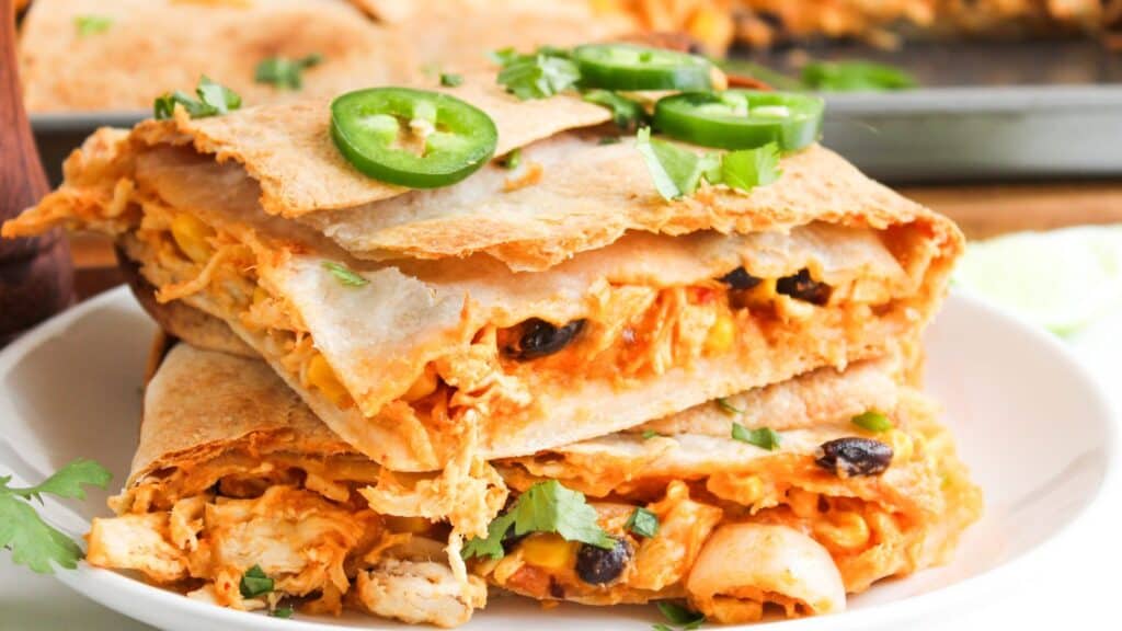 Close-up of stacked quesadillas filled with chicken, cheese, and black beans, garnished with sliced jalapeños and cilantro, on a white plate.