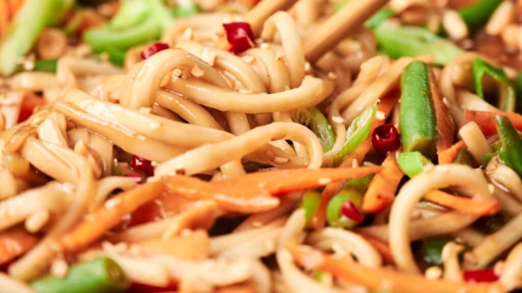 Close-up of stir-fried noodles with sliced carrots, green beans, and red chili peppers, garnished with sesame seeds. Chopsticks are seen in use.