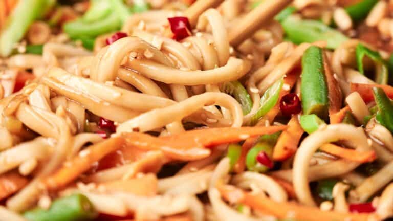 Close-up of stir-fried noodles with sliced carrots, green beans, and red chili peppers, garnished with sesame seeds. Chopsticks are seen in use.