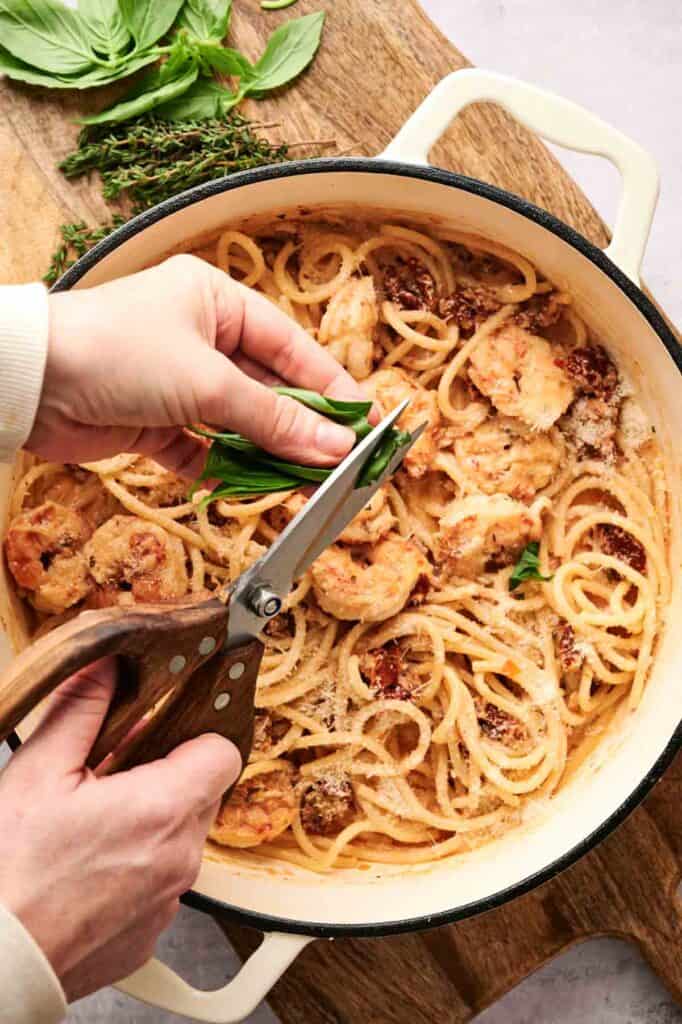 A hand expertly wields scissors to trim herbs over a luscious pot of creamy spaghetti, perfectly complemented with succulent chicken pieces, crafting an experience that could rival the captivating flavors of marry me shrimp pasta.
