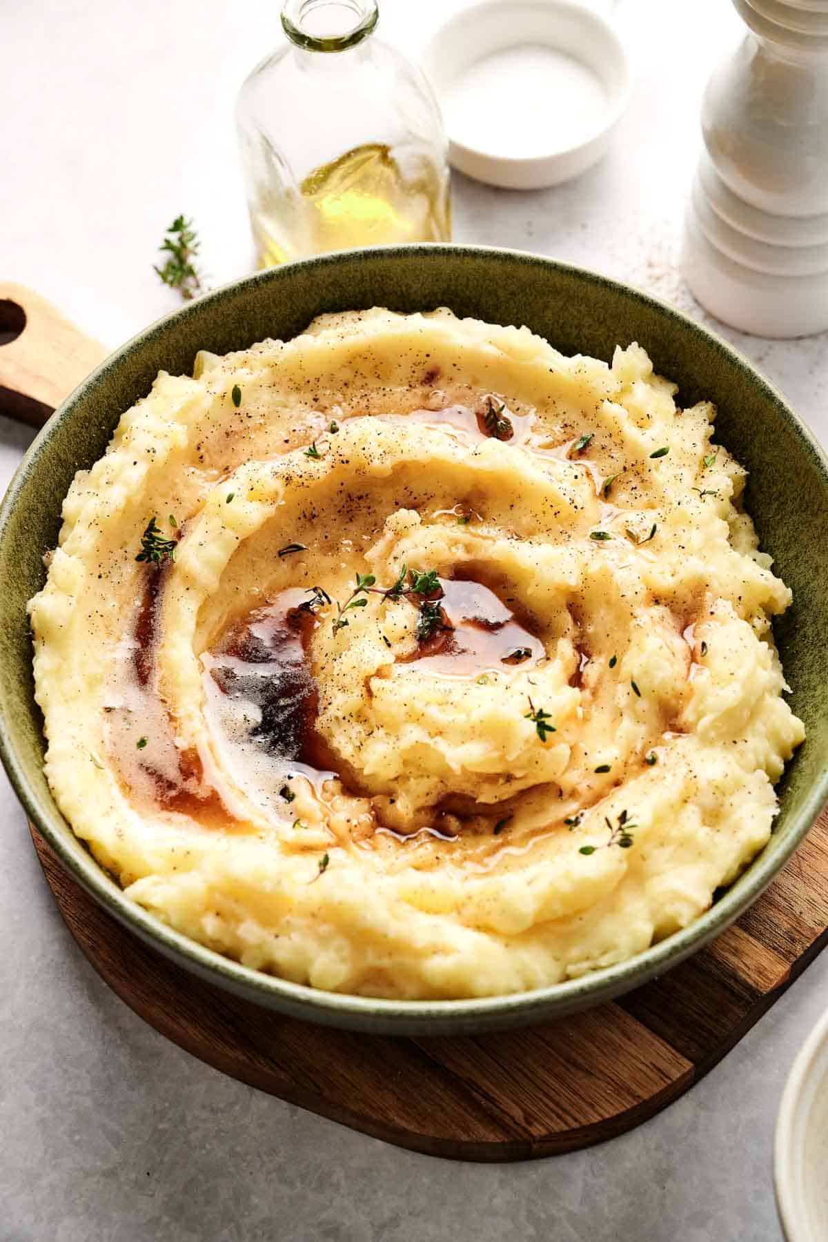 A green bowl brimming with creamy air fryer mashed potatoes, seasoned with butter, herbs, and pepper, sits gracefully on a wooden board. In the background, a bottle of oil and various seasonings await their turn.