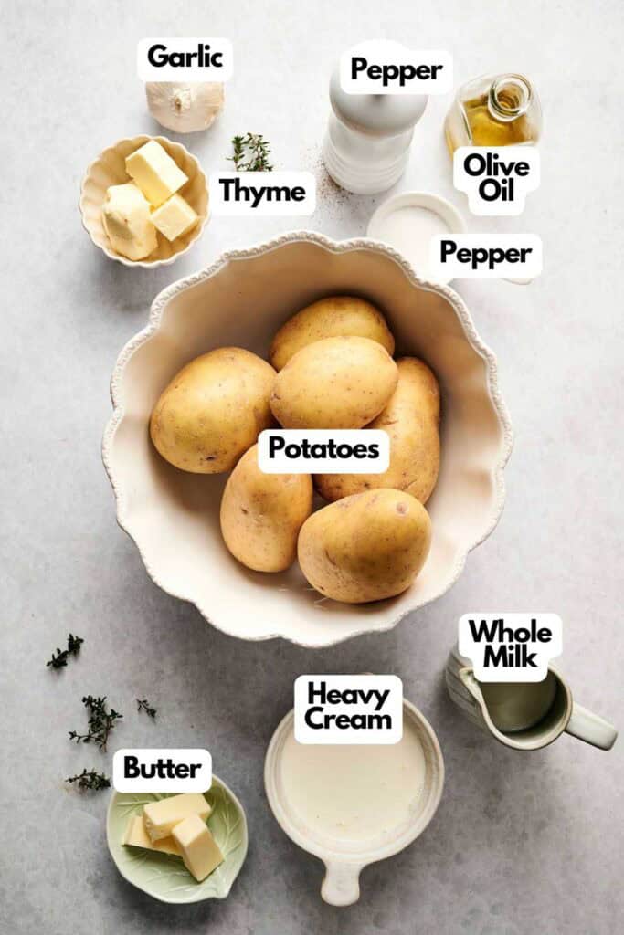 Ingredients for air fryer mashed potatoes are arranged on a surface, featuring creamy potatoes, fragrant garlic, fresh thyme, butter, rich heavy cream, whole milk, olive oil, and a sprinkle of pepper&mdash;all neatly labeled for easy prep.