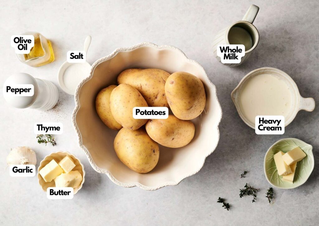Assorted ingredients for Air Fryer Mashed Potatoes, including potatoes in a bowl, butter, garlic, thyme, pepper, olive oil, salt, whole milk, and heavy cream, arranged on a kitchen counter.