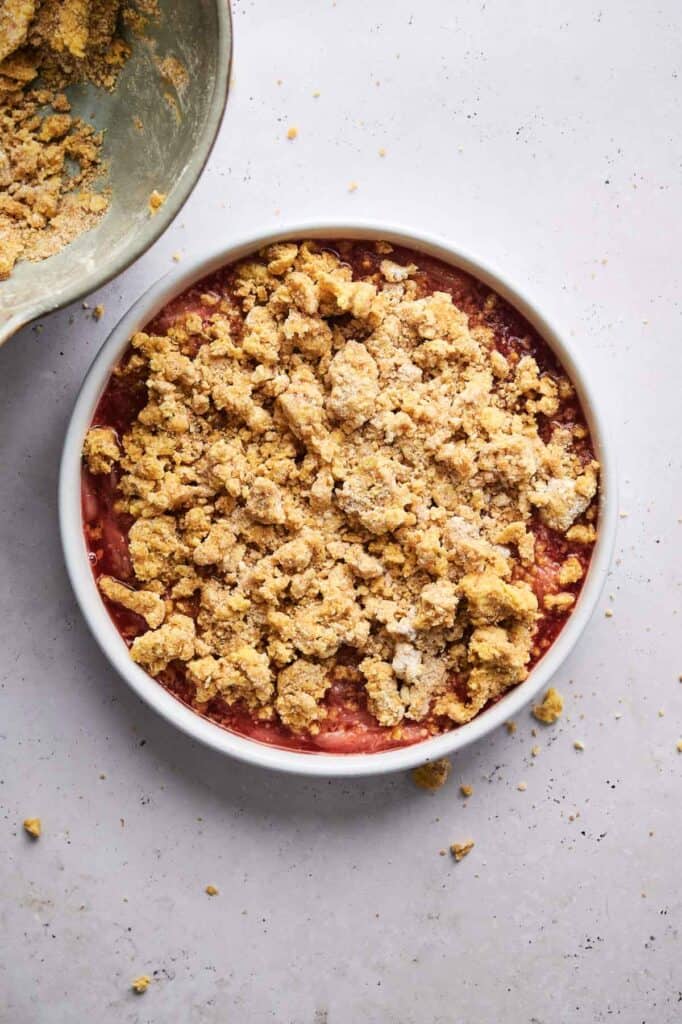 A round baking dish brimming with a rhubarb crisp mixture is topped with a golden-brown crumble. Loose crumbs are scattered around on a light surface.