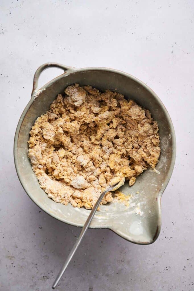 A mixing bowl with a crumbly flour and butter mixture, ready to transform into a delicious rhubarb crisp, sits alongside a spoon on a light surface.