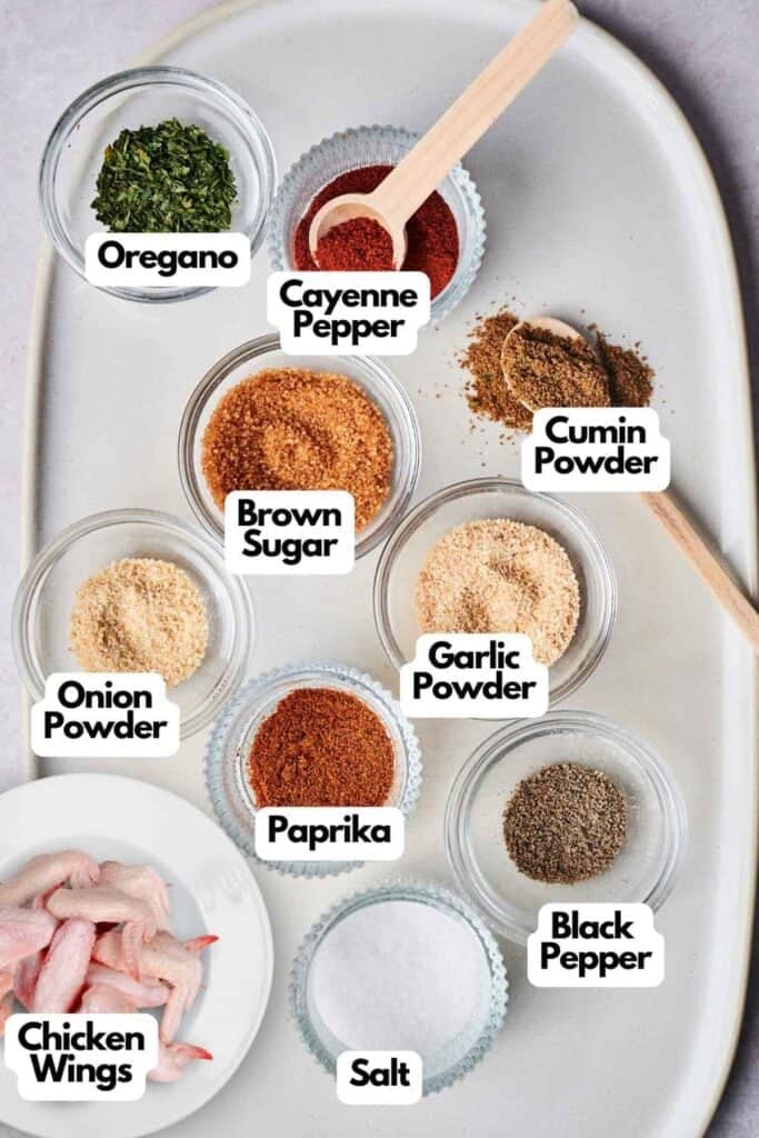 Ingredients for seasoning Wingstop Louisiana Rub Wings include bowls of oregano, cayenne pepper, cumin powder, onion powder, garlic powder, paprika, black pepper, brown sugar, and salt. Raw chicken wings shown.