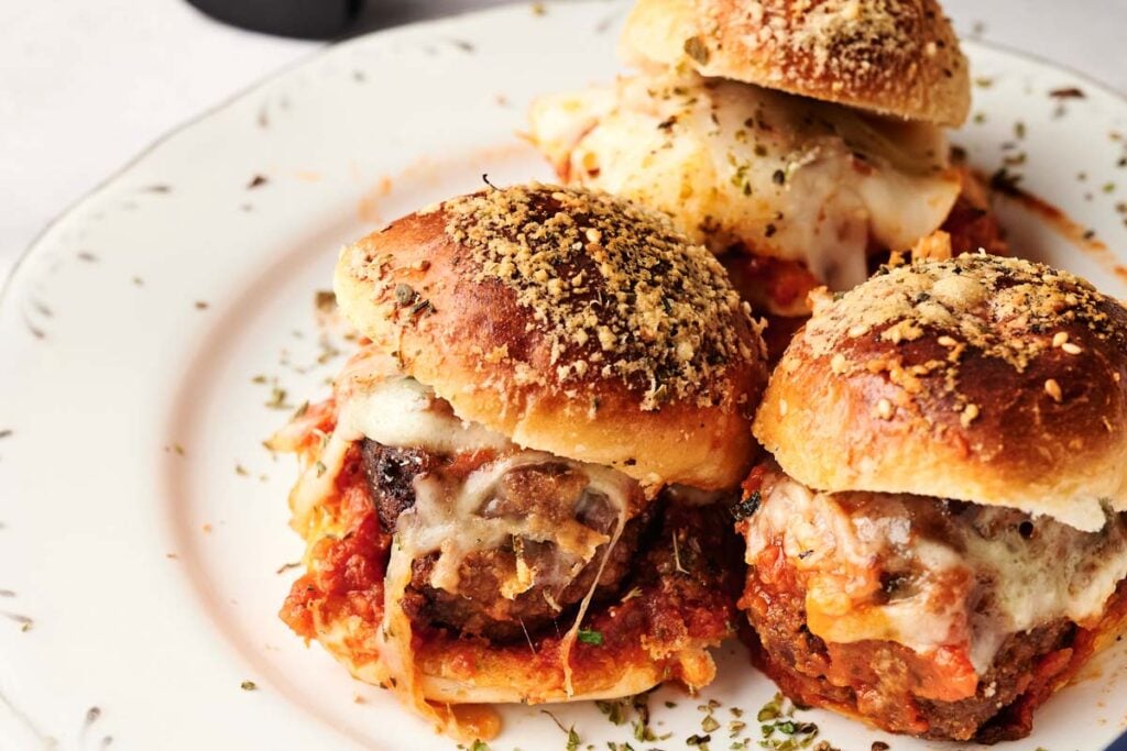 Three Air Fryer Meatball Sliders topped with melted cheese and herbs are arranged on a white plate, with tomato sauce visible beneath the buns.