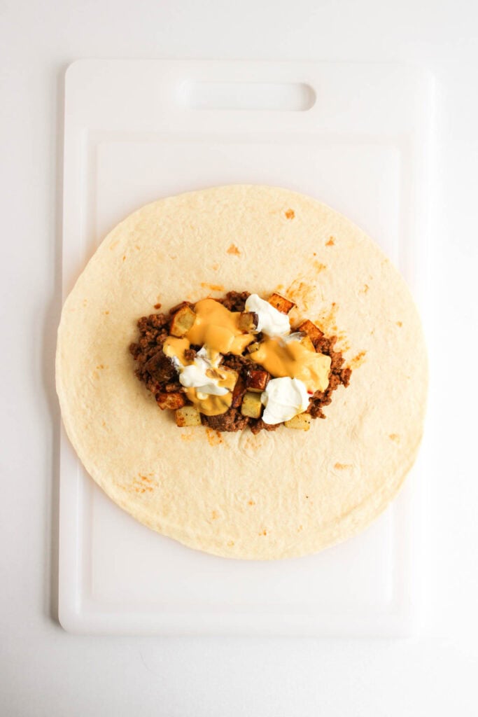 A flour tortilla on a white cutting board topped with ground beef, potatoes, cheese sauce, and sour cream&mdash;just like a Beef and Potato Burrito Taco Bell Copycat.