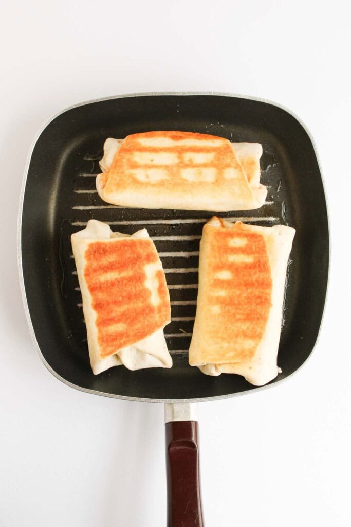 Three Beef and Potato Burrito Taco Bell Copycat burritos with golden-brown grill marks are cooking on a square grill pan against a white background.