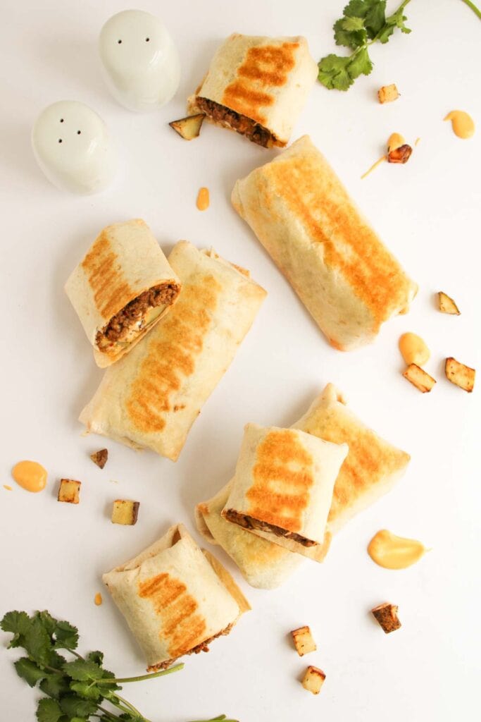 Grilled Beef and Potato Burrito Taco Bell Copycat burritos with visible grill marks are arranged on a white surface, surrounded by scattered cheese sauce, cilantro, and small tofu cubes. Two salt and pepper shakers are present.