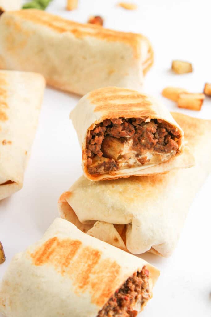 A close-up of several grilled Beef and Potato Burrito Taco Bell Copycat burritos, one cut in half to reveal a savory filling of ground beef, potatoes, and cheese, arranged on a white surface.