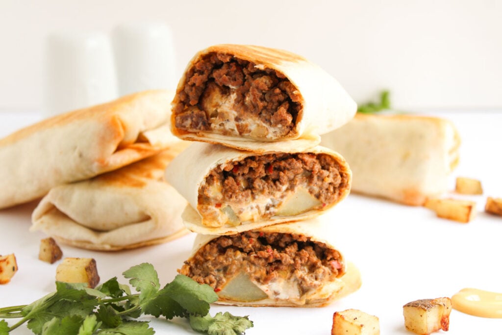 A stack of halved Beef and Potato Burrito Taco Bell Copycat burritos filled with ground beef and potatoes is shown on a white surface, with cubed potatoes and cilantro in the foreground.