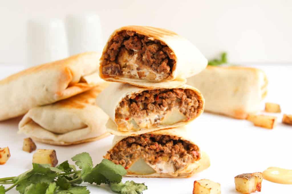 Three stacked Beef and Potato Burrito Taco Bell Copycat burritos are cut in half, showing their filling. Other whole burritos, diced potatoes, and cilantro are scattered around on a white surface.