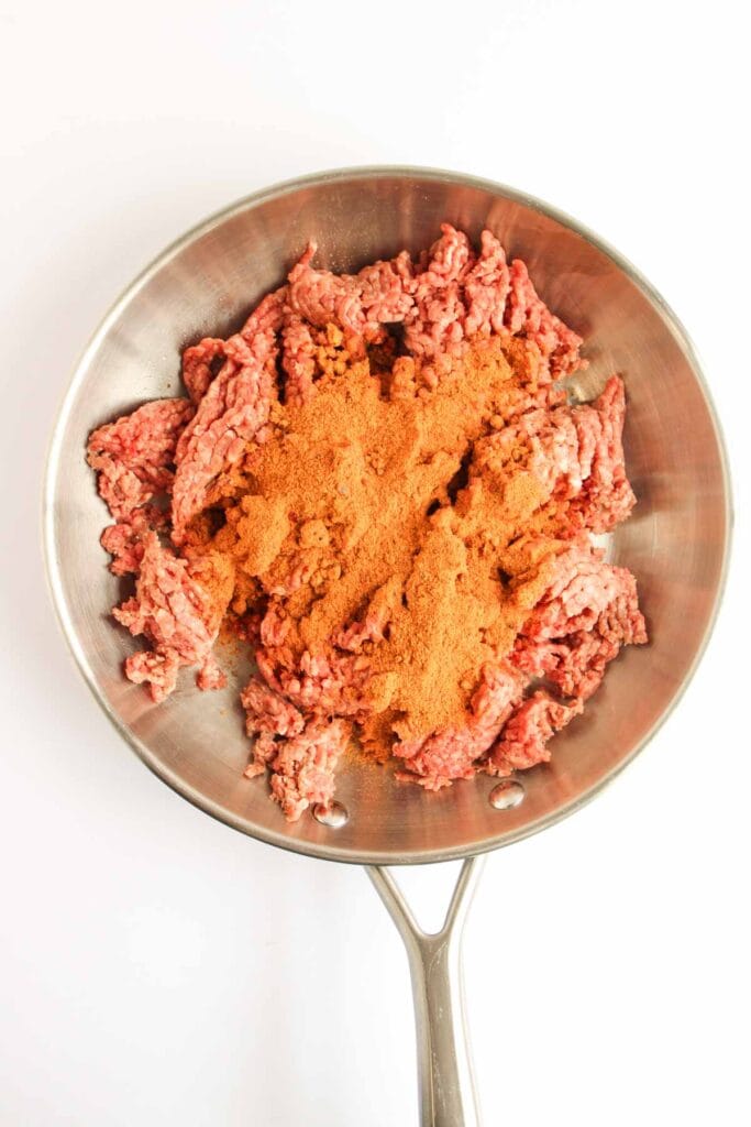 Ground beef topped with taco seasoning in a stainless steel skillet, perfect for making a Beef and Potato Burrito Taco Bell Copycat, photographed from above on a white background.