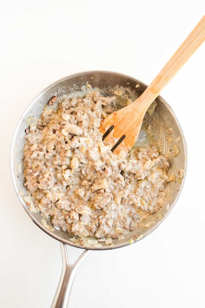 A stainless steel skillet filled with cooked ground meat, onions, and creamy sauce&mdash;perfect for making Big Mac Sloppy Joes&mdash;being stirred with a wooden spatula.
