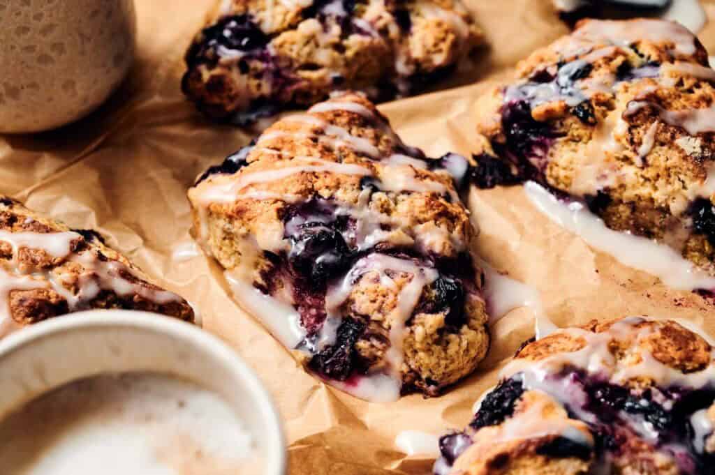 Close-up of blueberry scones with icing drizzled on top, arranged on brown parchment paper with a cup of coffee nearby&mdash;perfect for pairing with a slice of ice cream cake for an indulgent treat.