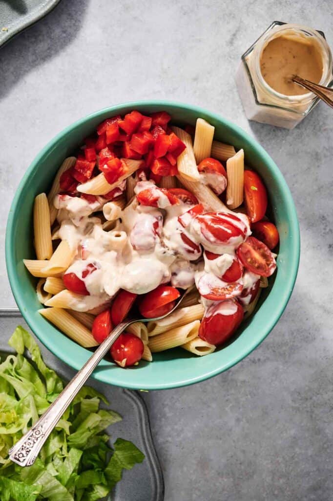 A bowl of penne pasta with cherry tomatoes, diced red bell pepper, and creamy sauce evokes the essence of a Chicken Caesar Pasta Salad. A container of sauce and a plate of lettuce are nearby on a gray surface.