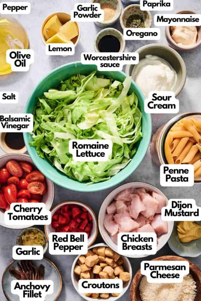 An assortment of ingredients in bowls for a Chicken Caesar Pasta Salad, featuring romaine lettuce, pasta, chicken breasts, cherry tomatoes, red bell pepper, croutons, and condiments like mayonnaise, mustard, and spices.