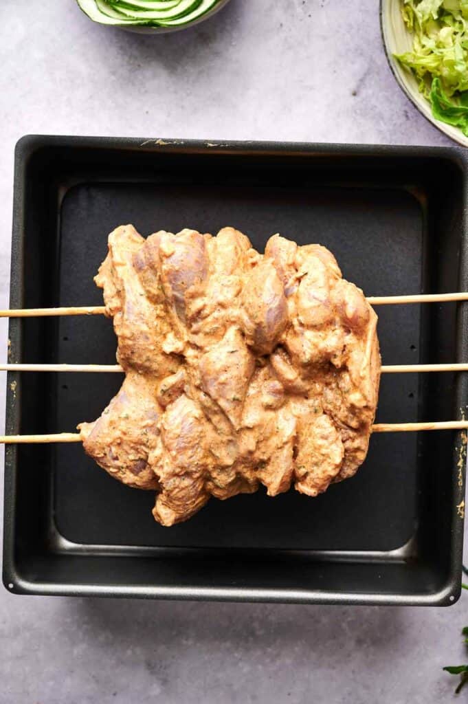 Raw marinated chicken pieces threaded on skewers placed over a black baking tray.
