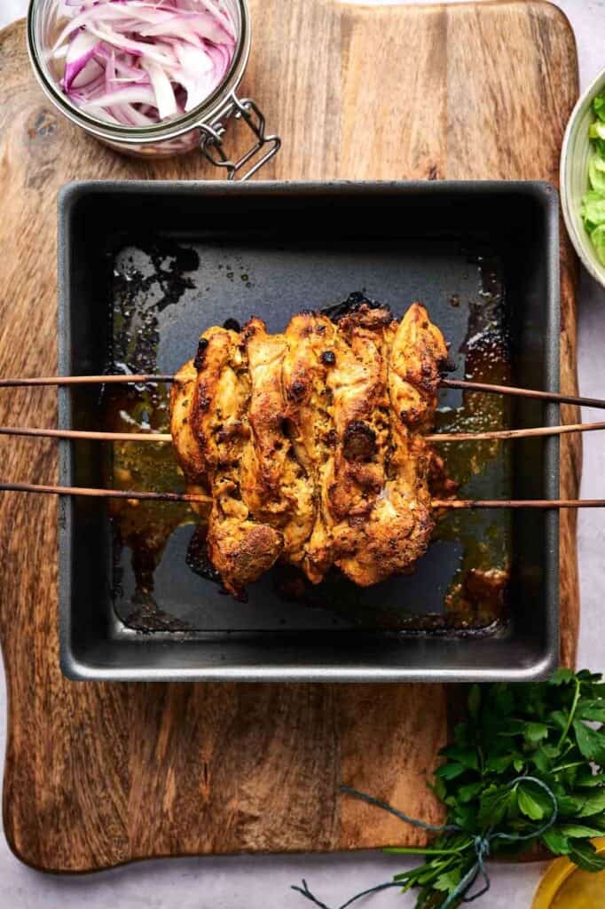 Grilled marinated chicken skewered in a square pan, placed on a wooden board. Nearby are sliced onions in a jar and green herbs.
