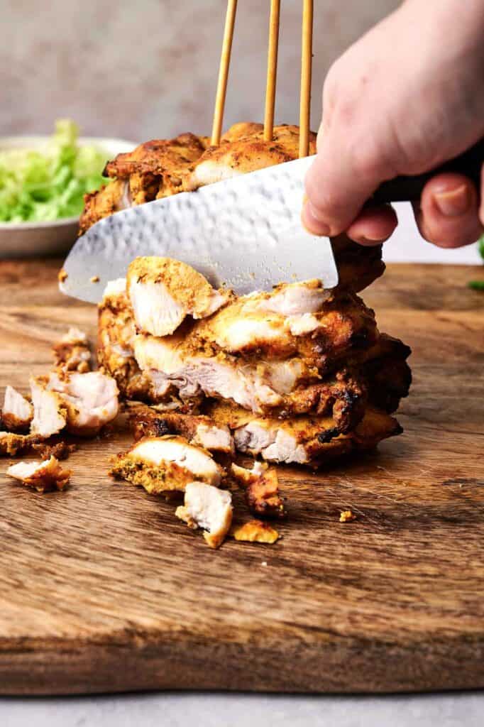 A hand slices cooked marinated chicken stacked on skewers on a wooden board, with a bowl of lettuce in the background.