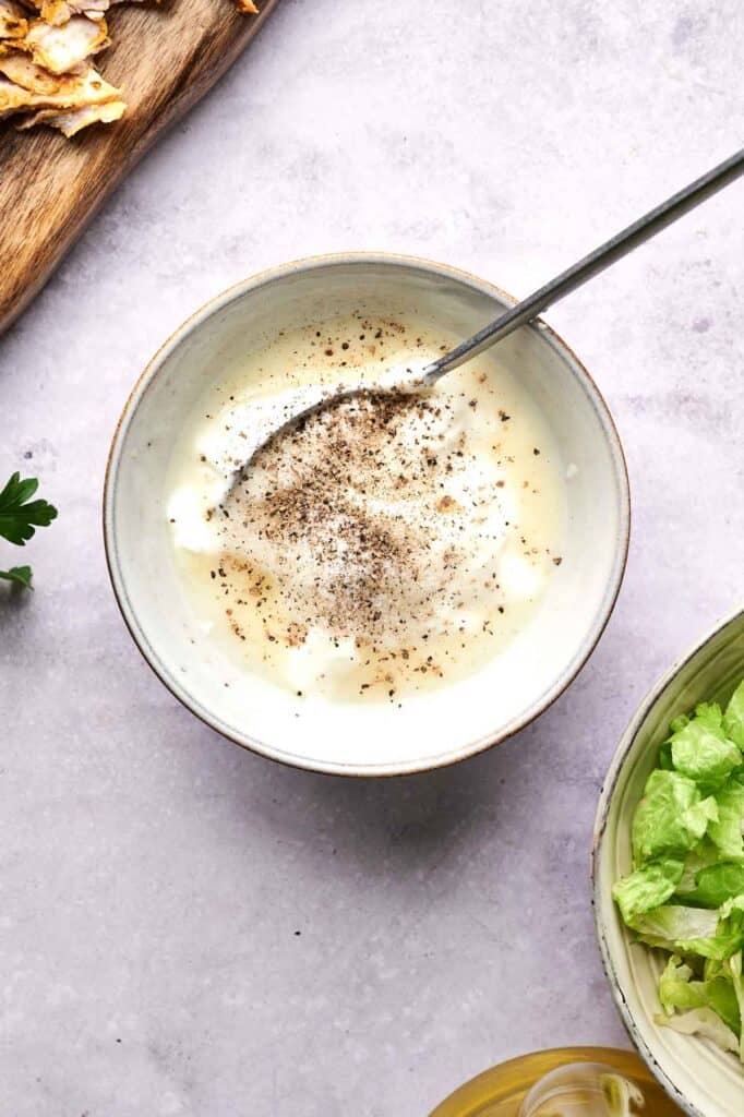 A bowl of creamy salad dressing with black pepper on top, next to lettuce and a wooden board with crackers.