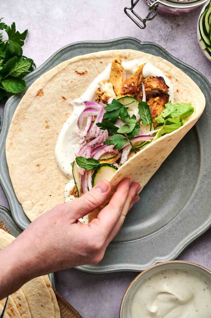 A hand holds a Chicken Shawarma flatbread wrap filled with grilled chicken, sliced onions, cucumbers, lettuce, fresh herbs, and white sauce on a gray plate.