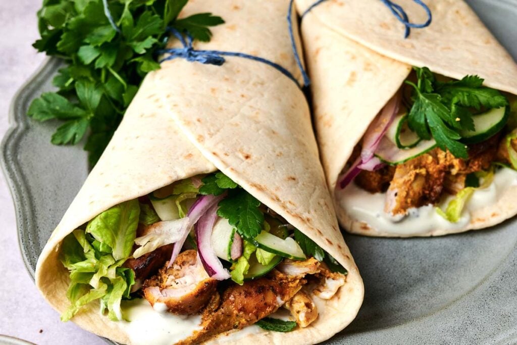 Two Chicken Shawarma wraps filled with lettuce, red onion, fresh parsley, and sauce, tied with blue string, served on a grey plate.